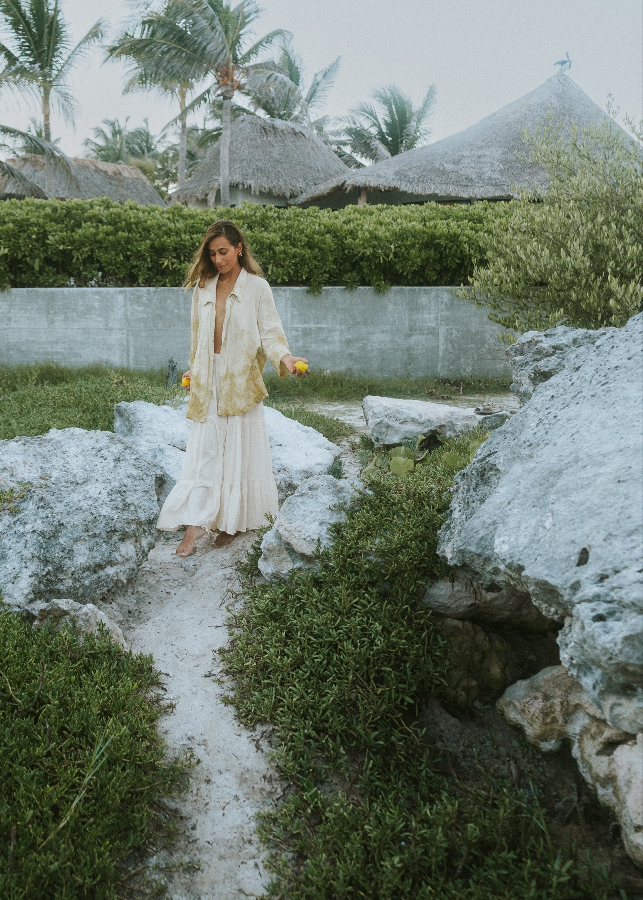 A woman in botanically dyed linen walks a limestone path between thatched palapas and low greenery.