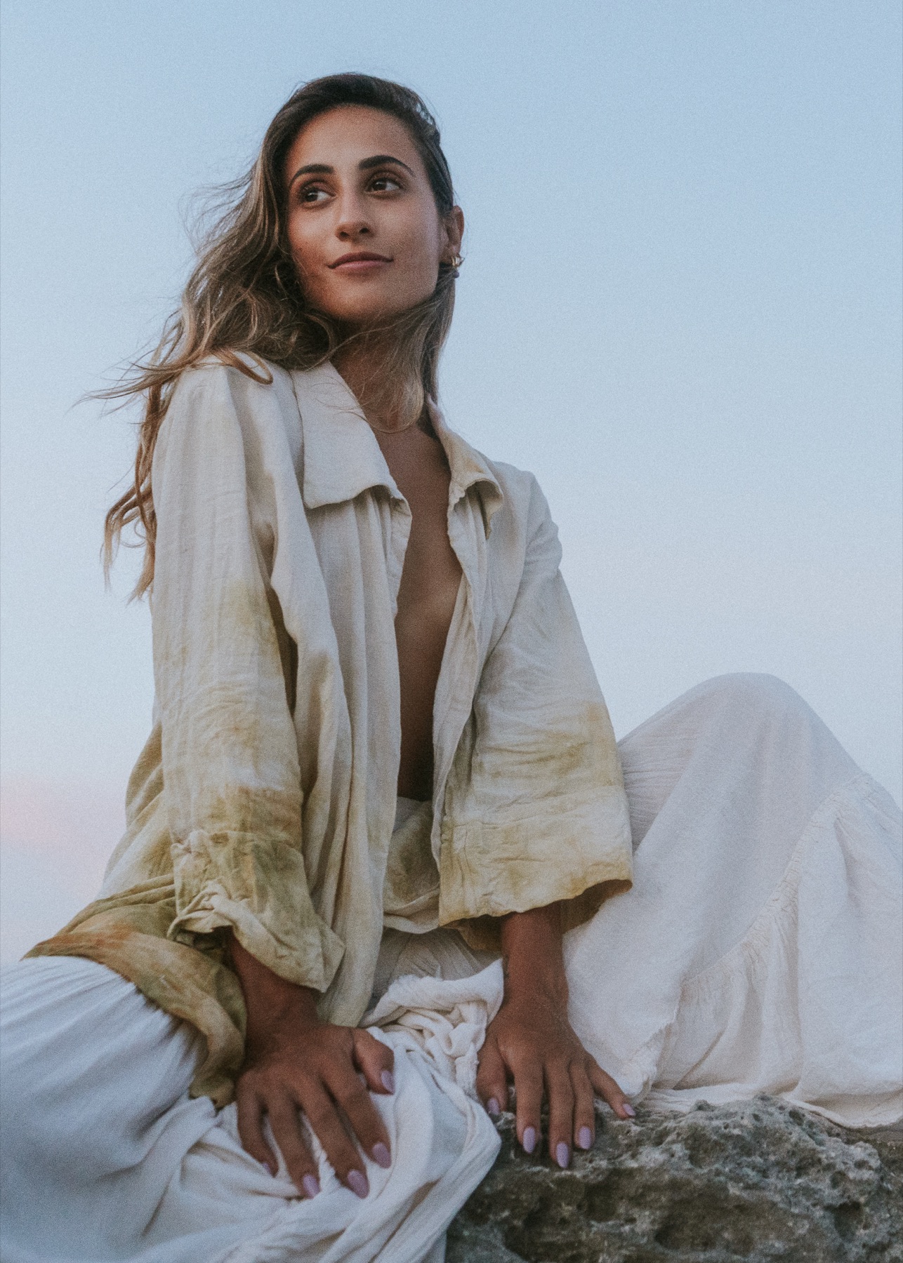 A woman in a botanically dyed jacket and white linen sits on rock against a pale dusk sky.