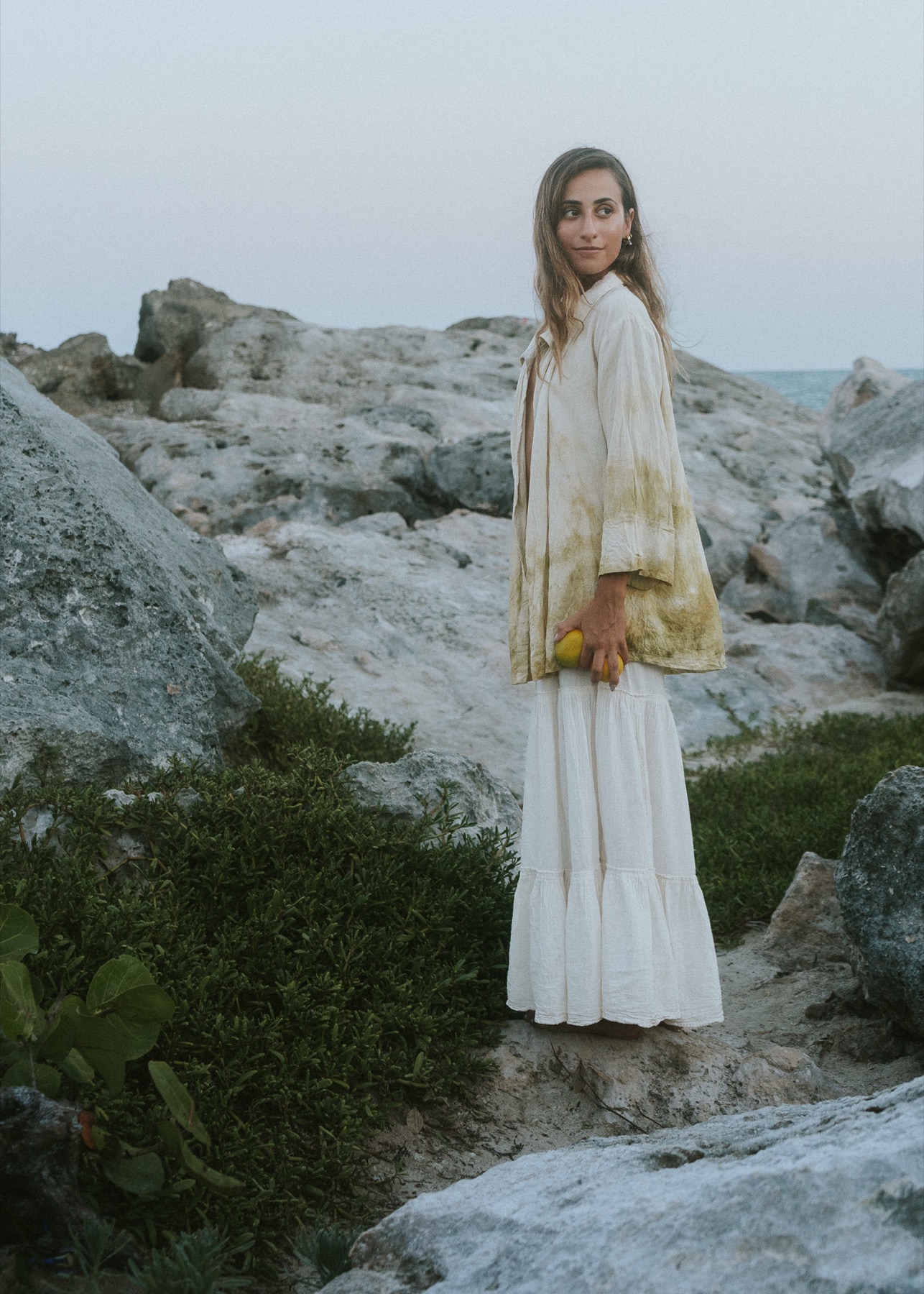 A woman in botanically dyed linen stands on limestone rocks holding a lemon, sea behind her.