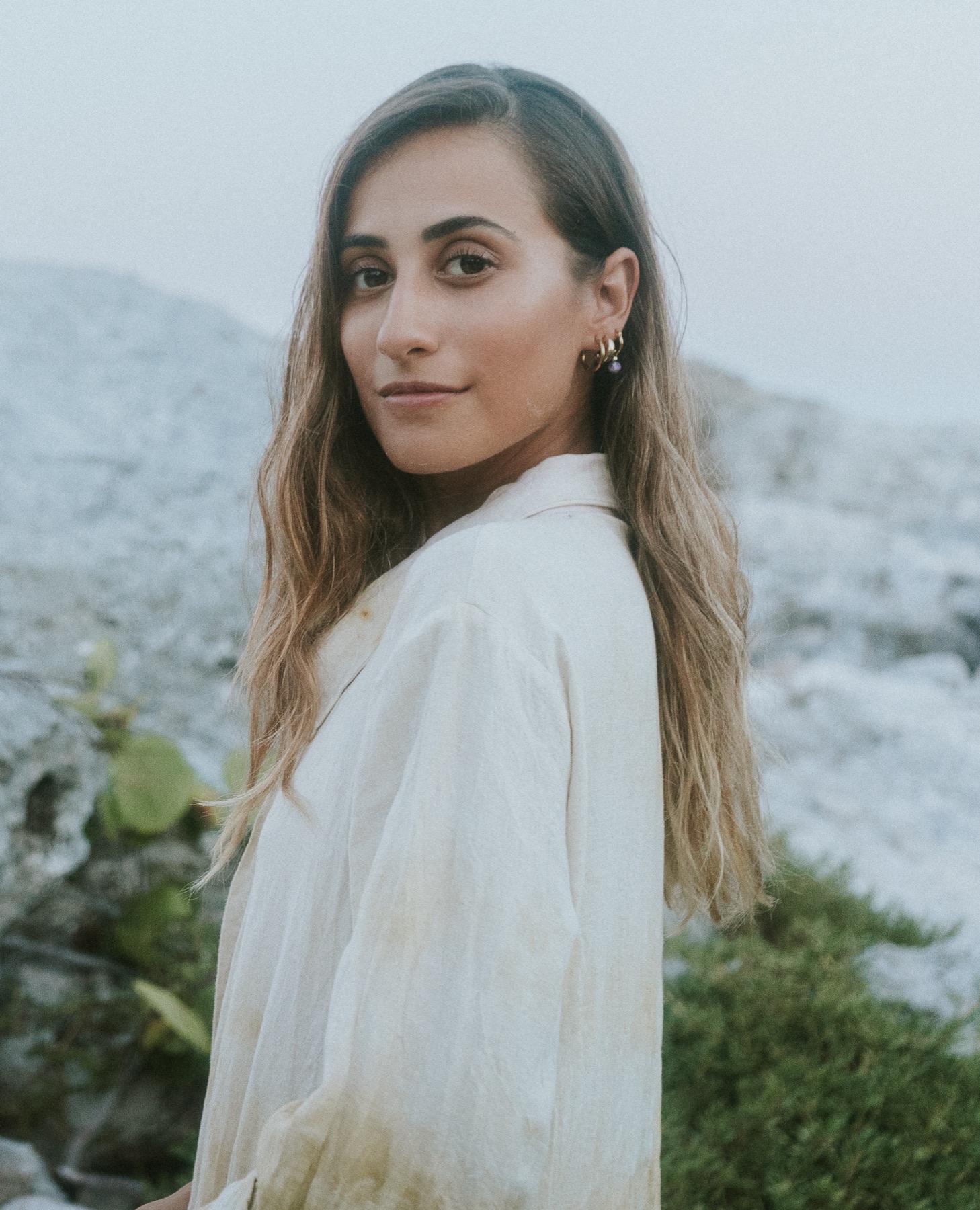 A woman in a botanically dyed linen jacket turns to camera, limestone and low scrub behind her.