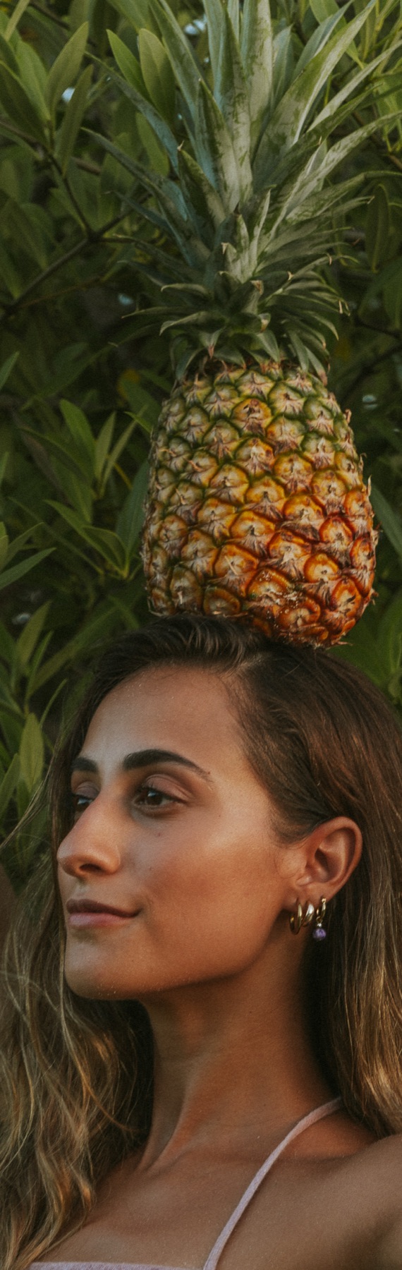 A woman stands beneath a pineapple balanced above her head against tropical foliage.