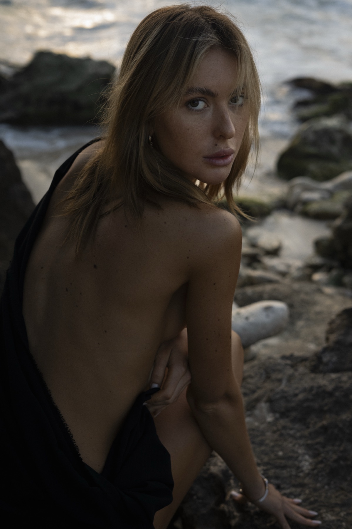 A woman in a black backless dress looks over her shoulder, crouched on wet rocks at the shoreline.
