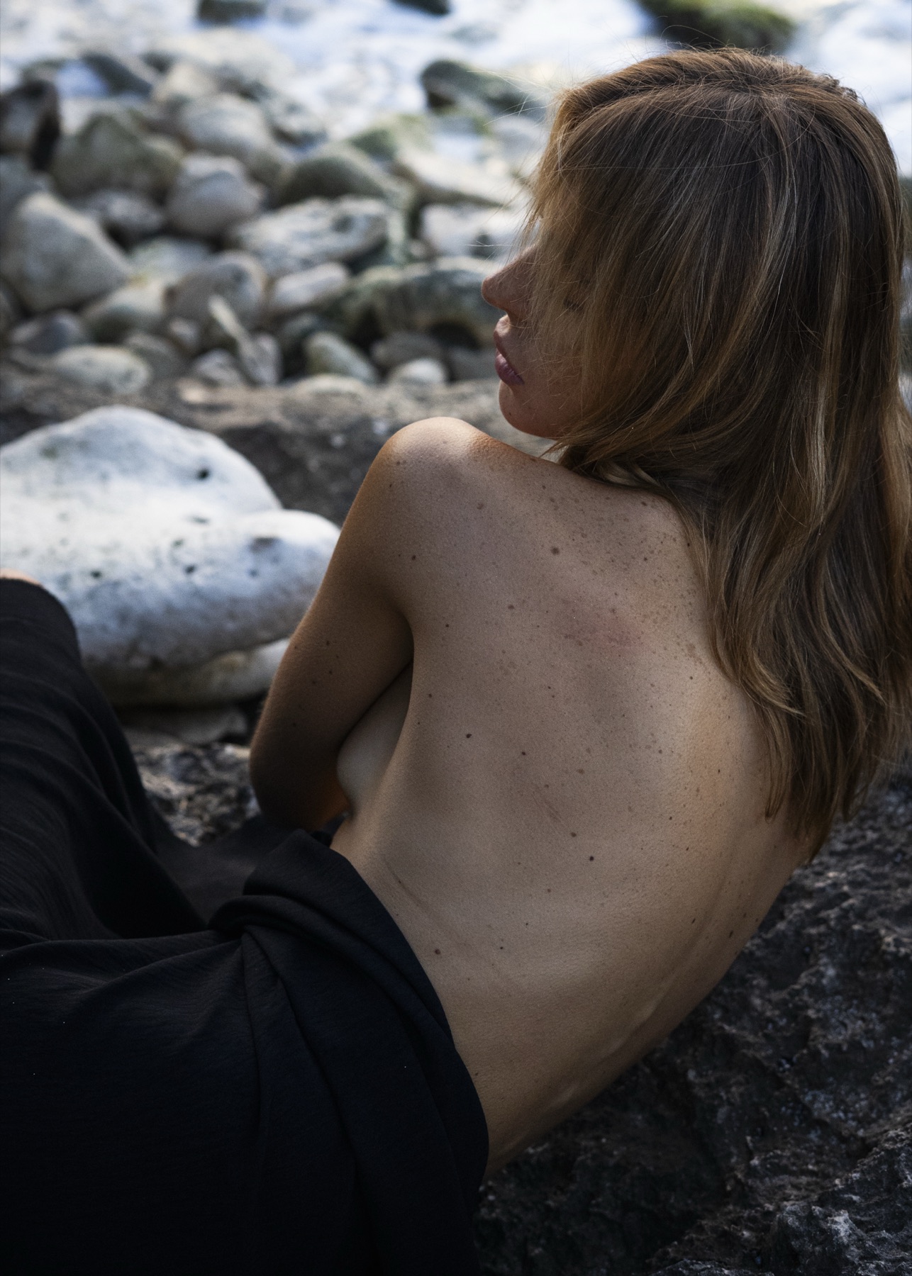 A woman sits bare-backed on rock among pale pebbles, freckled shoulders turned to the sea.