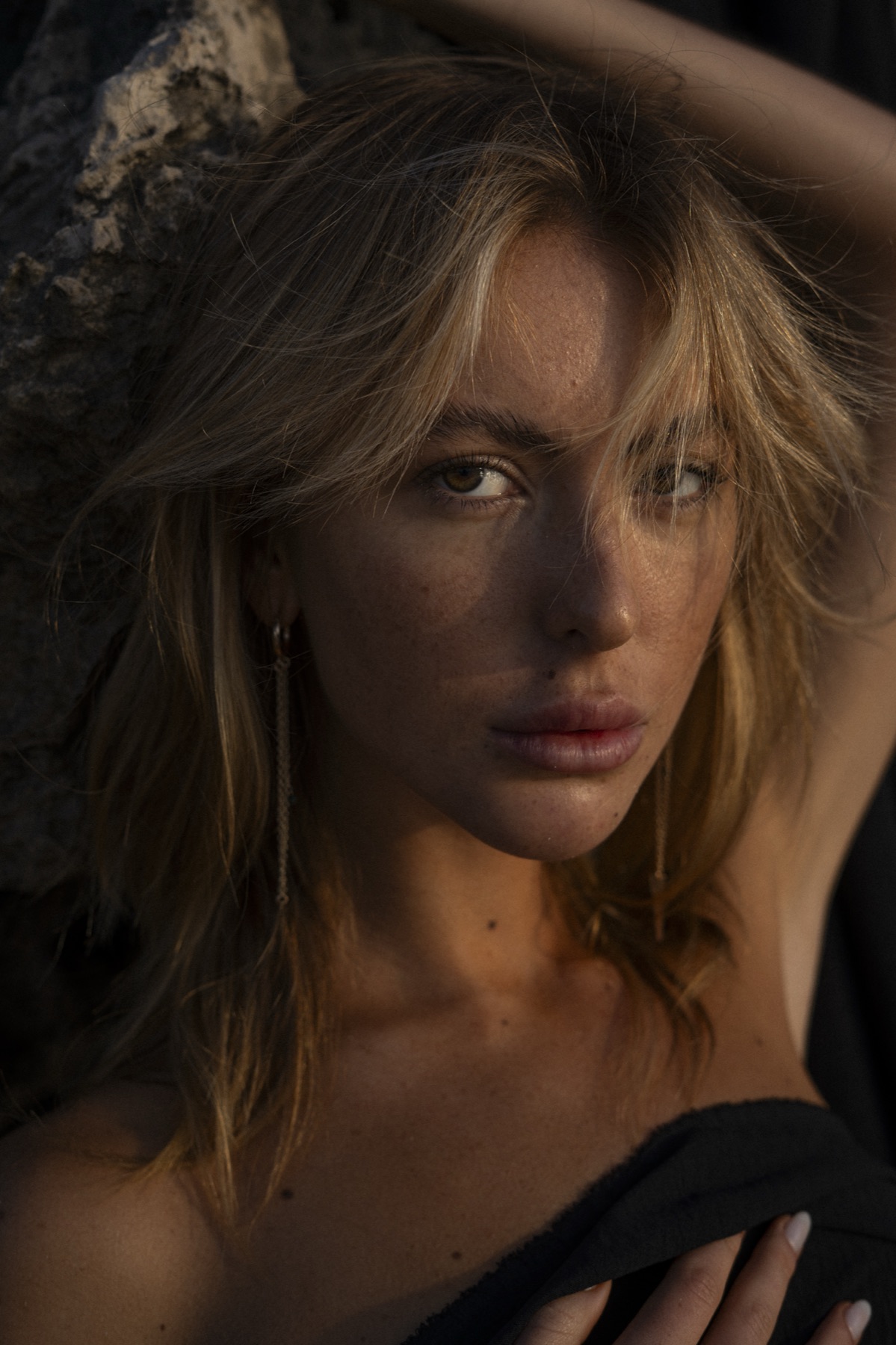 Close portrait of a woman with sun-bleached hair and a long chain earring against limestone rock.
