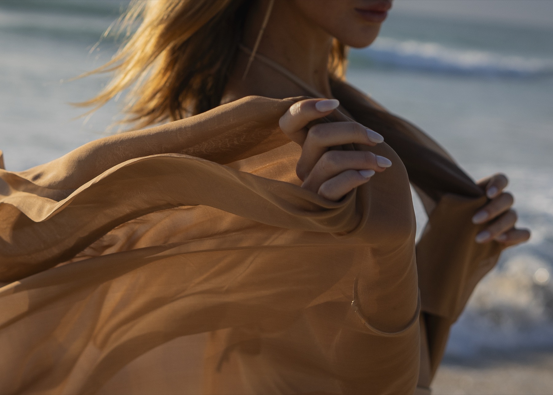 Detail of hands drawing a caramel silk scarf across a bare shoulder, sea and sunlight behind.