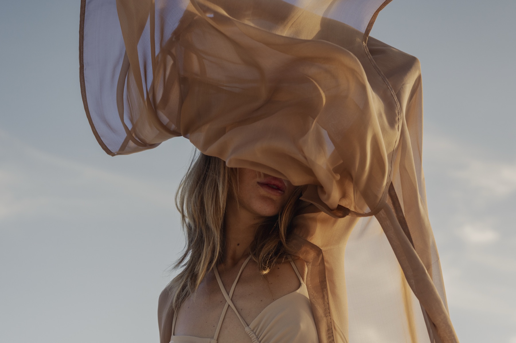 A sheer caramel scarf blows across a woman's face and raised arms against a pale sky.