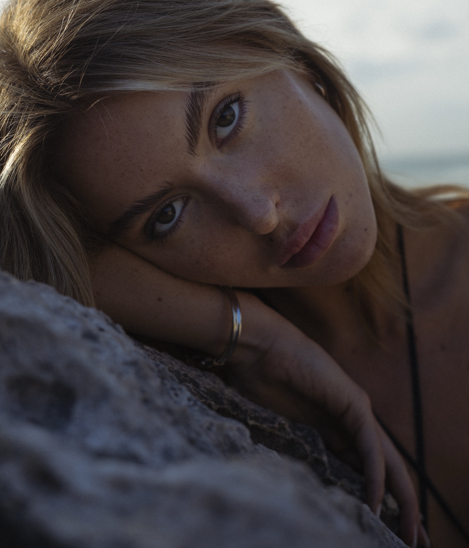 A woman rests her cheek and hand on limestone rock, freckled face turned to the camera at dusk.