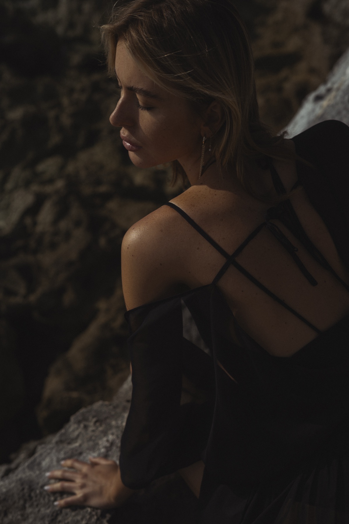 A woman in a black dress with crossed back straps looks down, turned away against a rock wall.