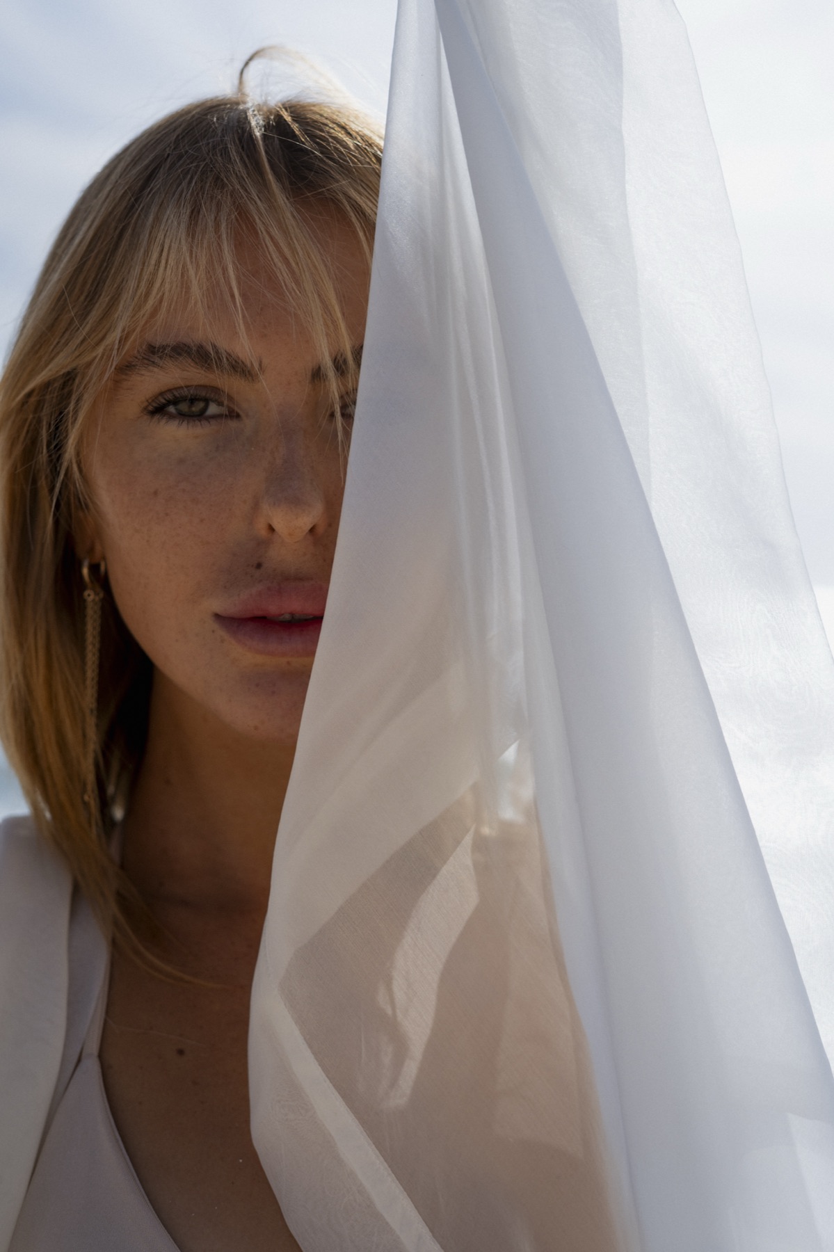 A woman's face half hidden behind a white sheet lifted against the sky.