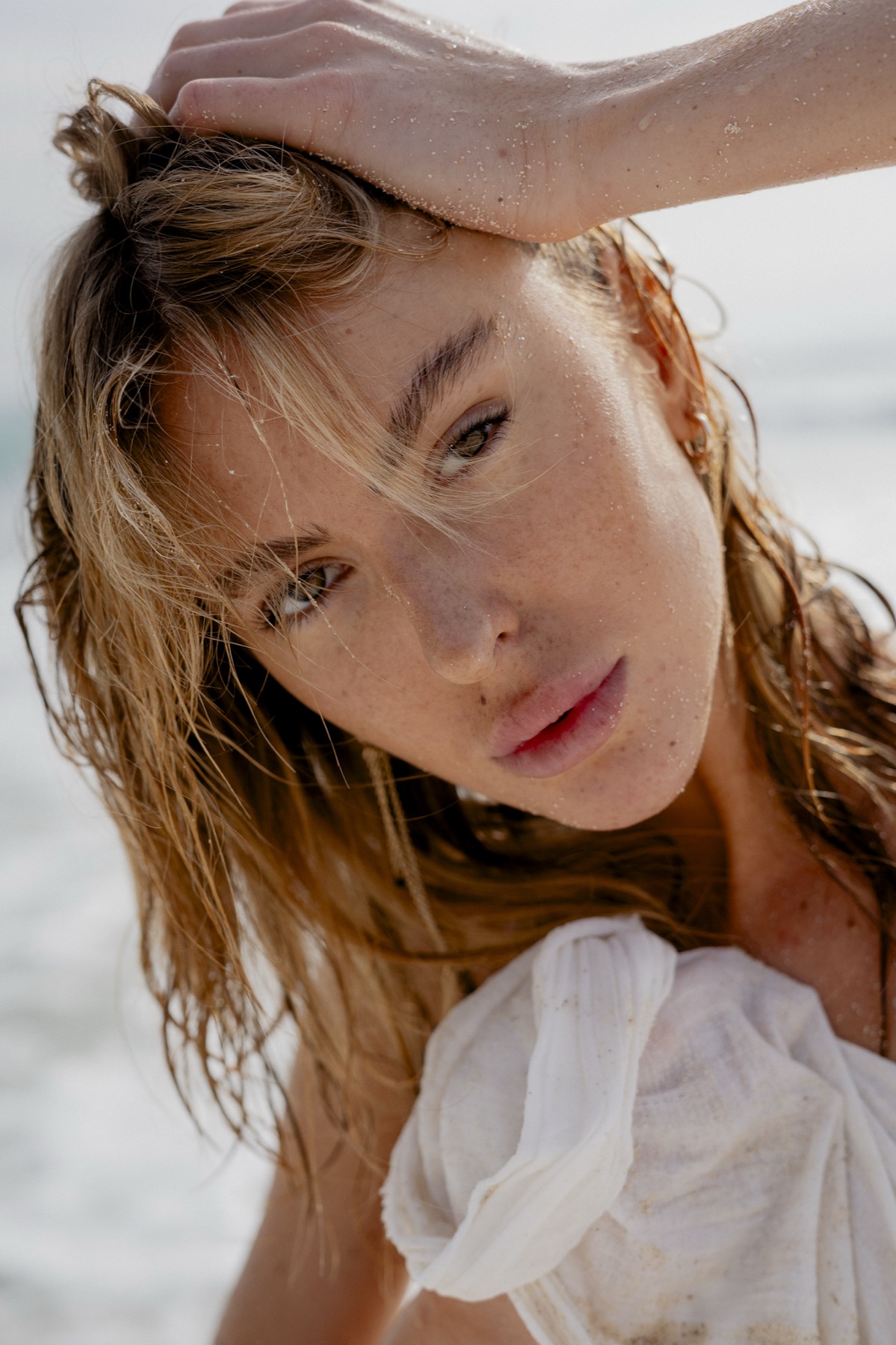 A woman with wet salt-flecked hair pushes it back from her face on the beach, white cotton at her shoulder.