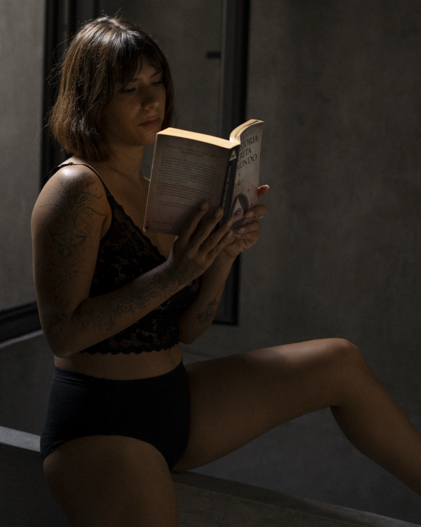 A woman in black lace sits on a concrete ledge reading a Spanish paperback held in both hands.