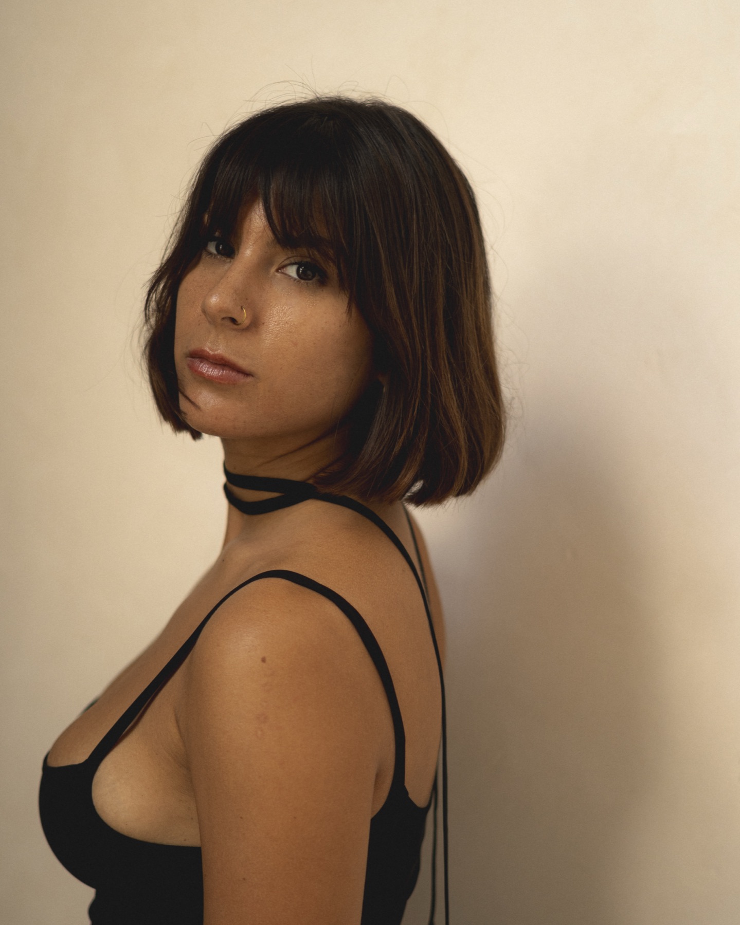 Profile portrait of a woman with a blunt bob in a black slip dress against a warm cream wall.