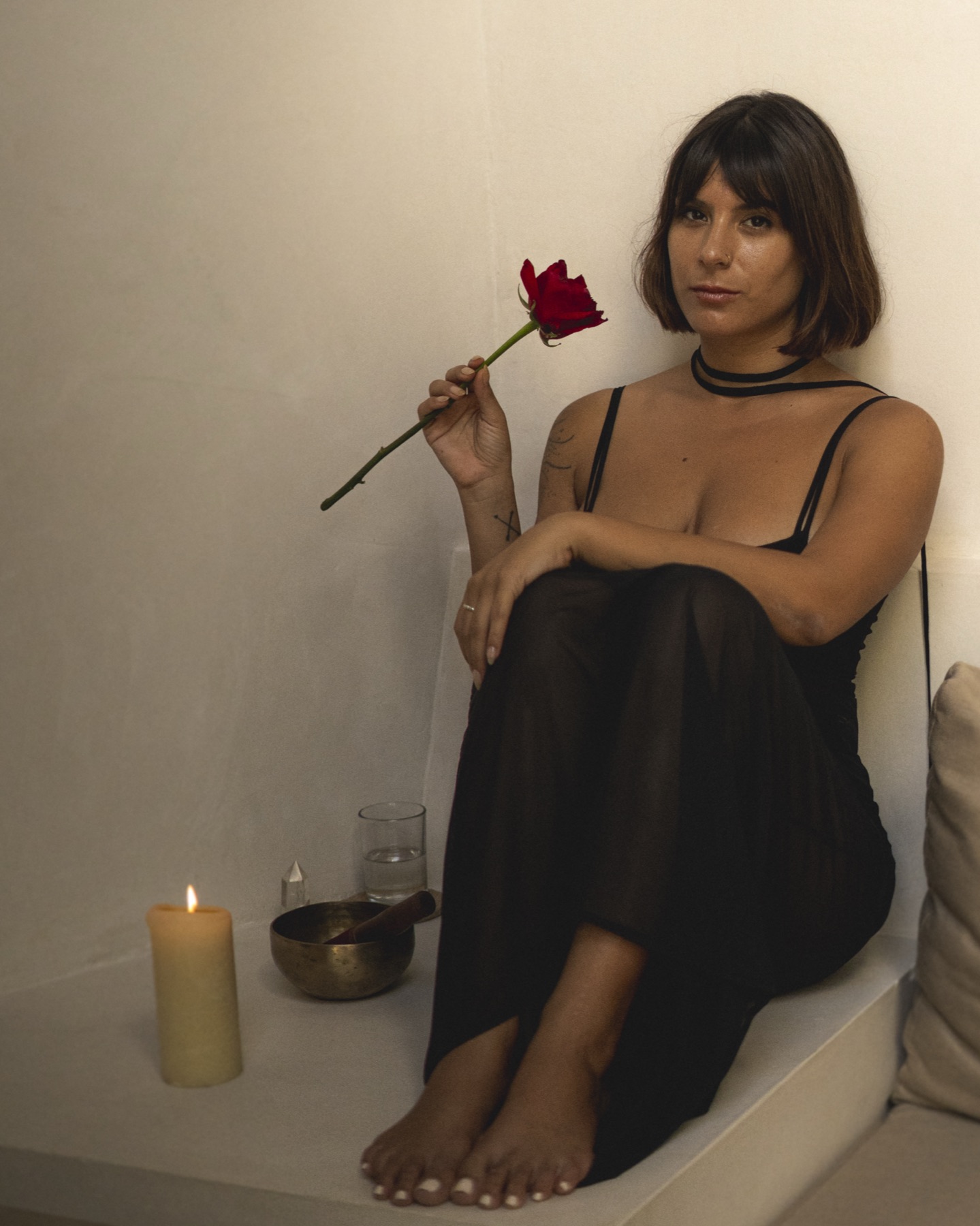 A woman in a sheer black dress sits in a whitewashed nook holding a single red rose, candle and singing bowl beside her.