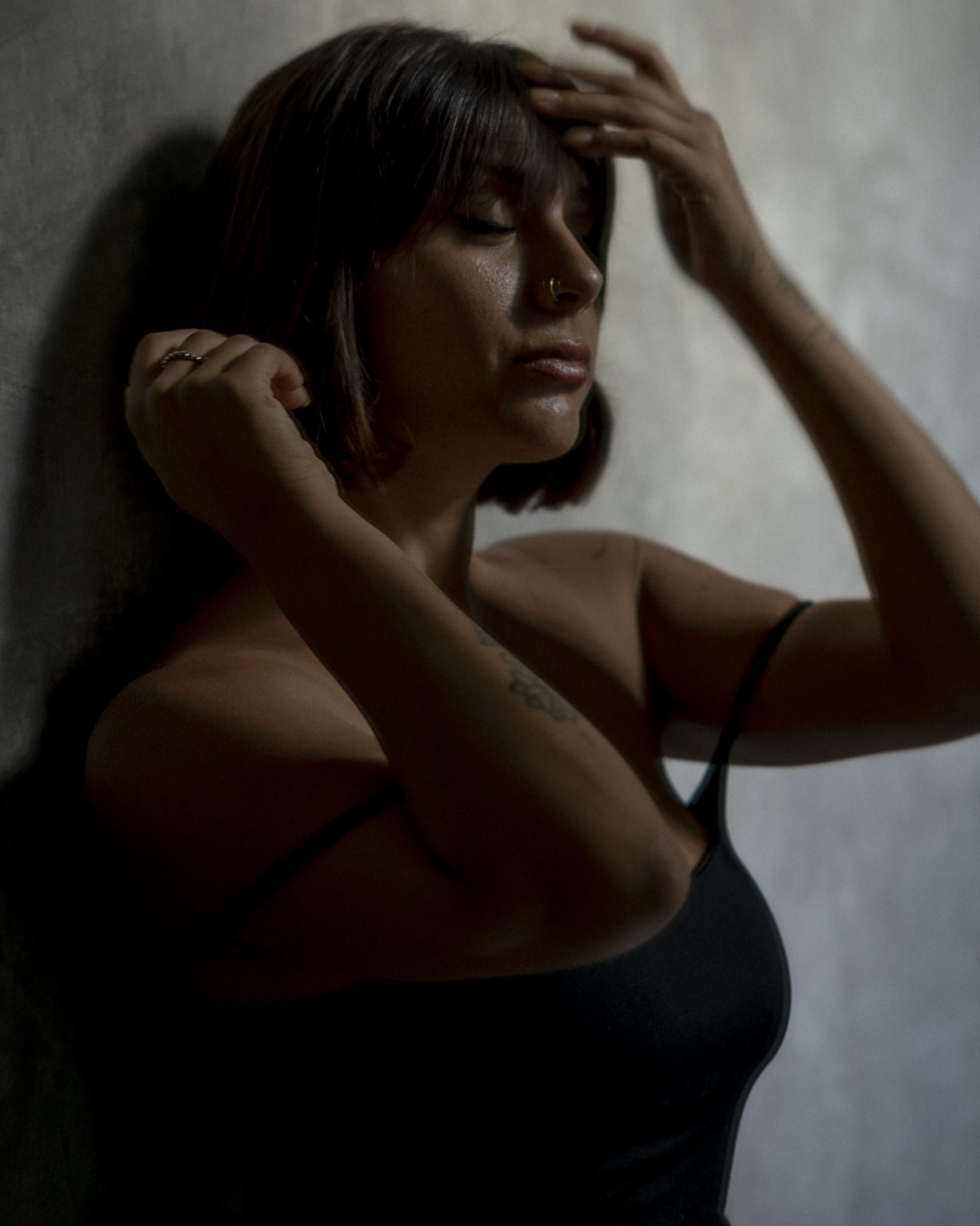 A woman in a black slip leans against a concrete wall with both hands raised to her hair, eyes closed.