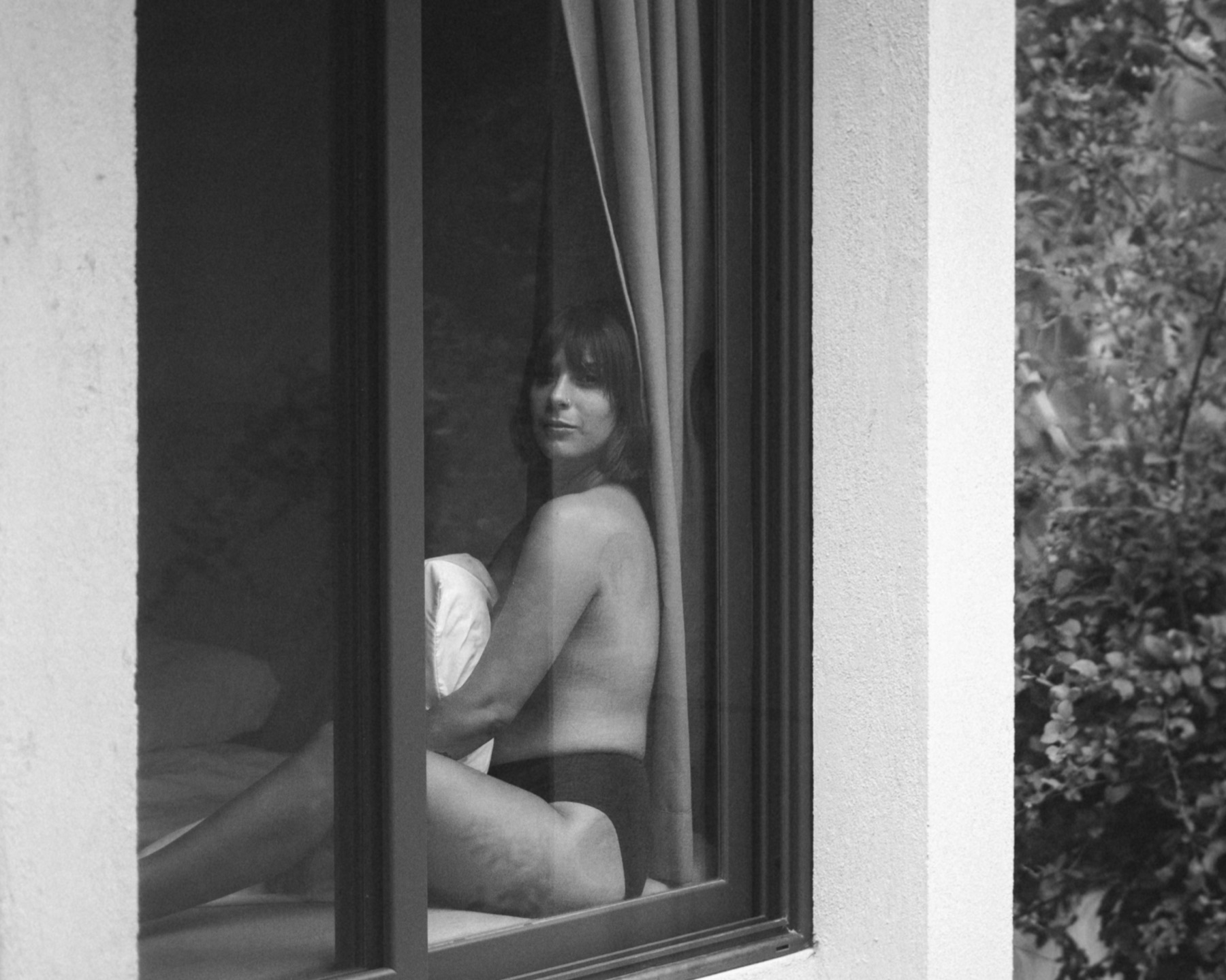 Black and white frame of a woman seen through an open window, sitting on a bed behind a curtain.