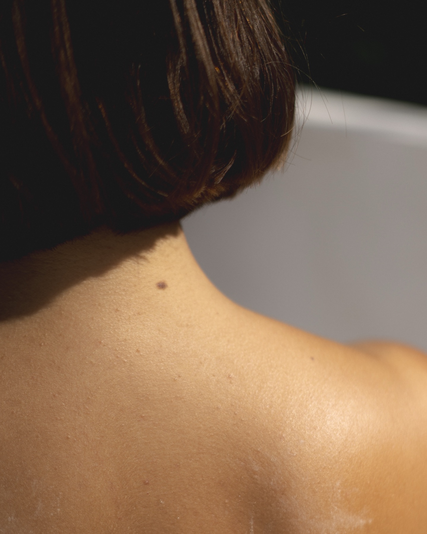 Close detail of a woman's shoulder and the nape of her neck under her dark bob, warm side light.