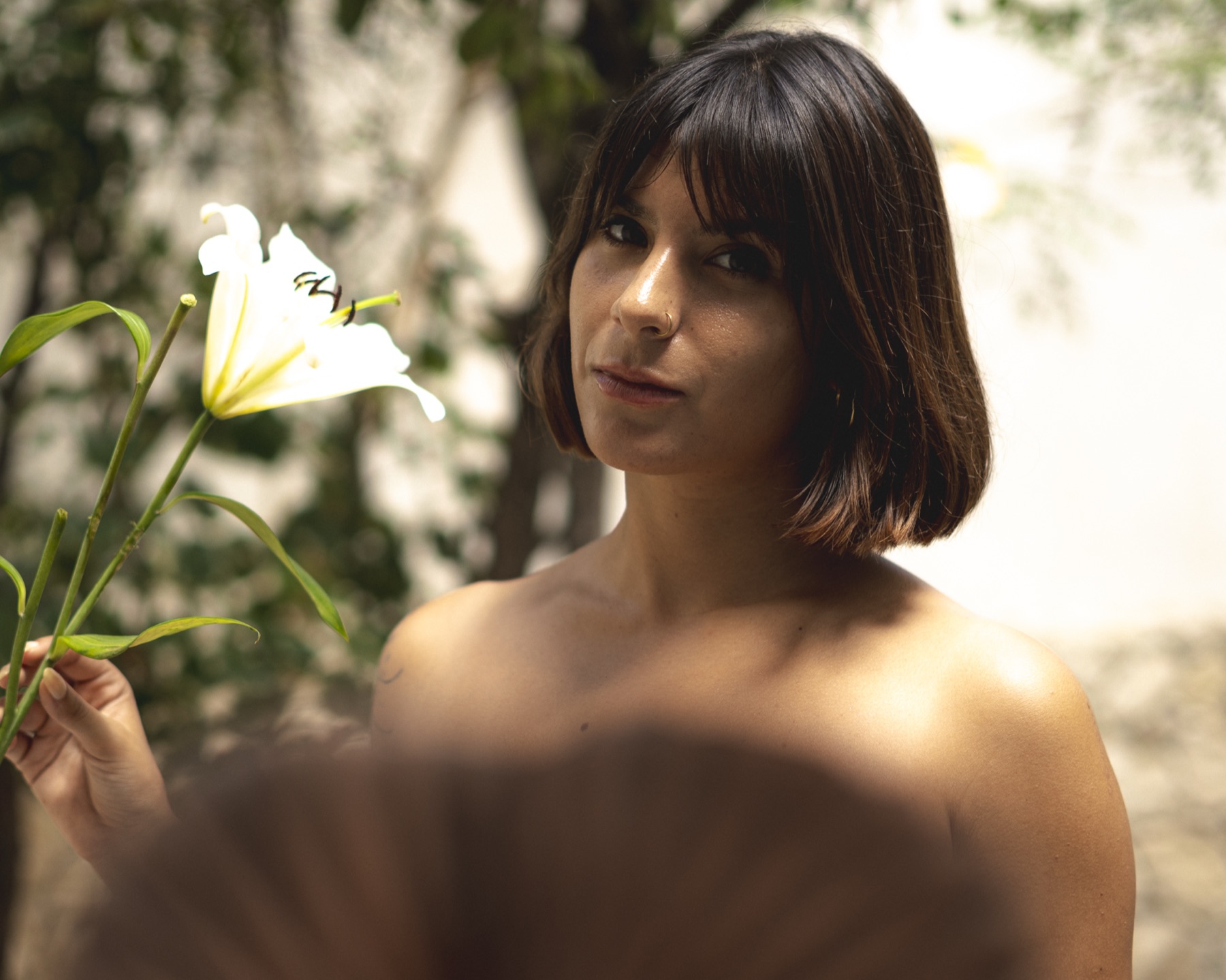 A woman looks to camera beside a white lily, tropical foliage and a sunlit wall behind her.