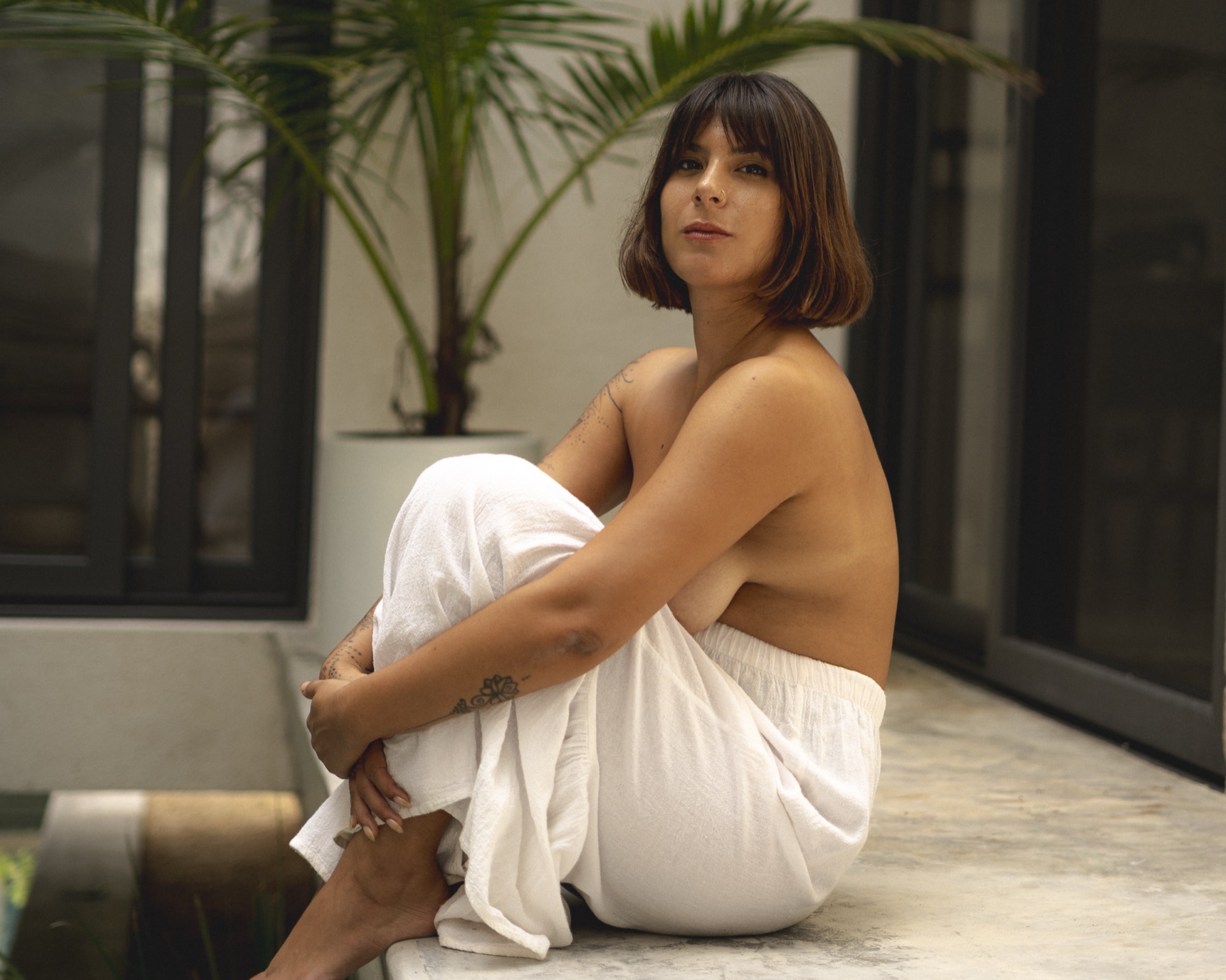 A woman sits on a terrace ledge wrapped in white linen with a potted palm behind her.